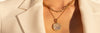 A person wearing a cream-colored blazer with layered gold necklaces, one featuring a paperclip chain and the other a round pendant with decorative accents.